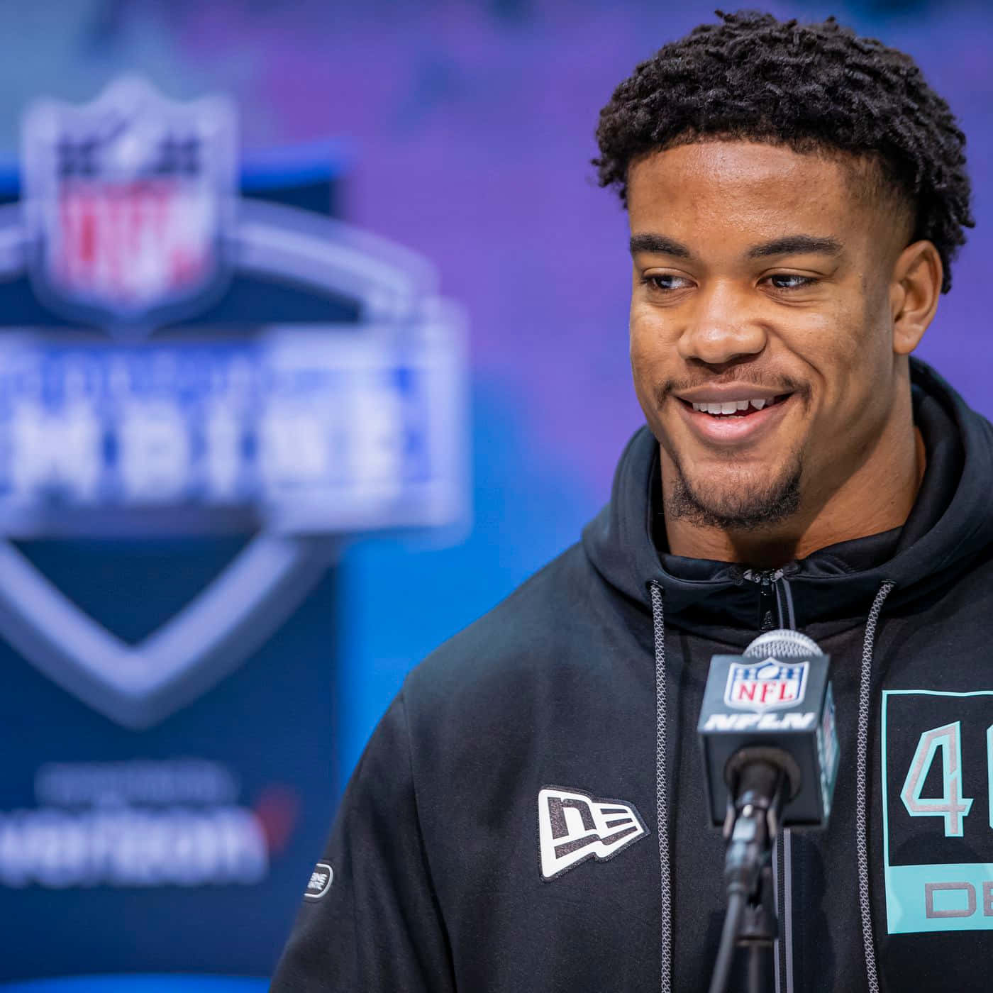  壁紙 — 一位微笑的NFL球員穿著黑色連帽衫，在NFL聯合試訓期間於講台上發言，背景有模糊的標誌。