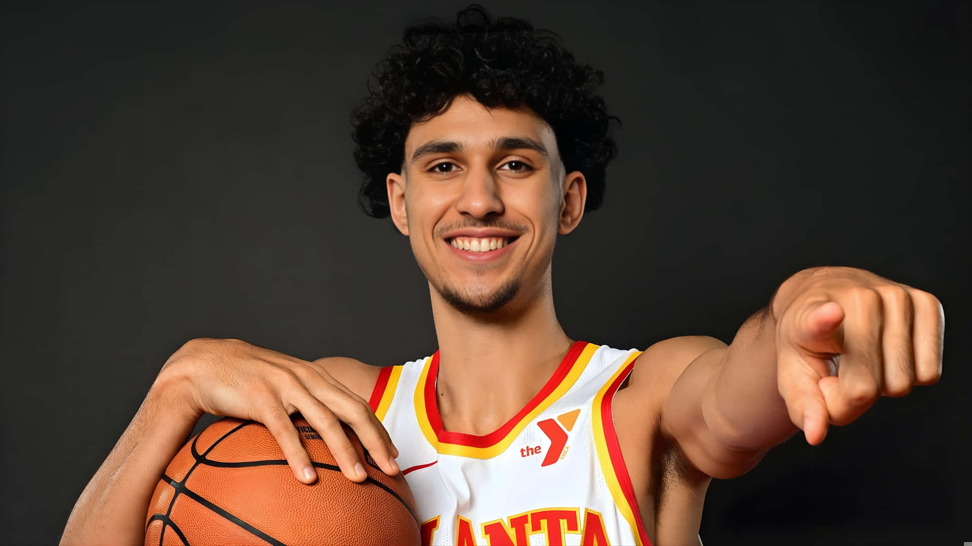  壁紙 — 一位微笑的年輕籃球運動員身穿白色亞特蘭大球衣，在深色工作室背景下自信地拿著球並指向鏡頭。