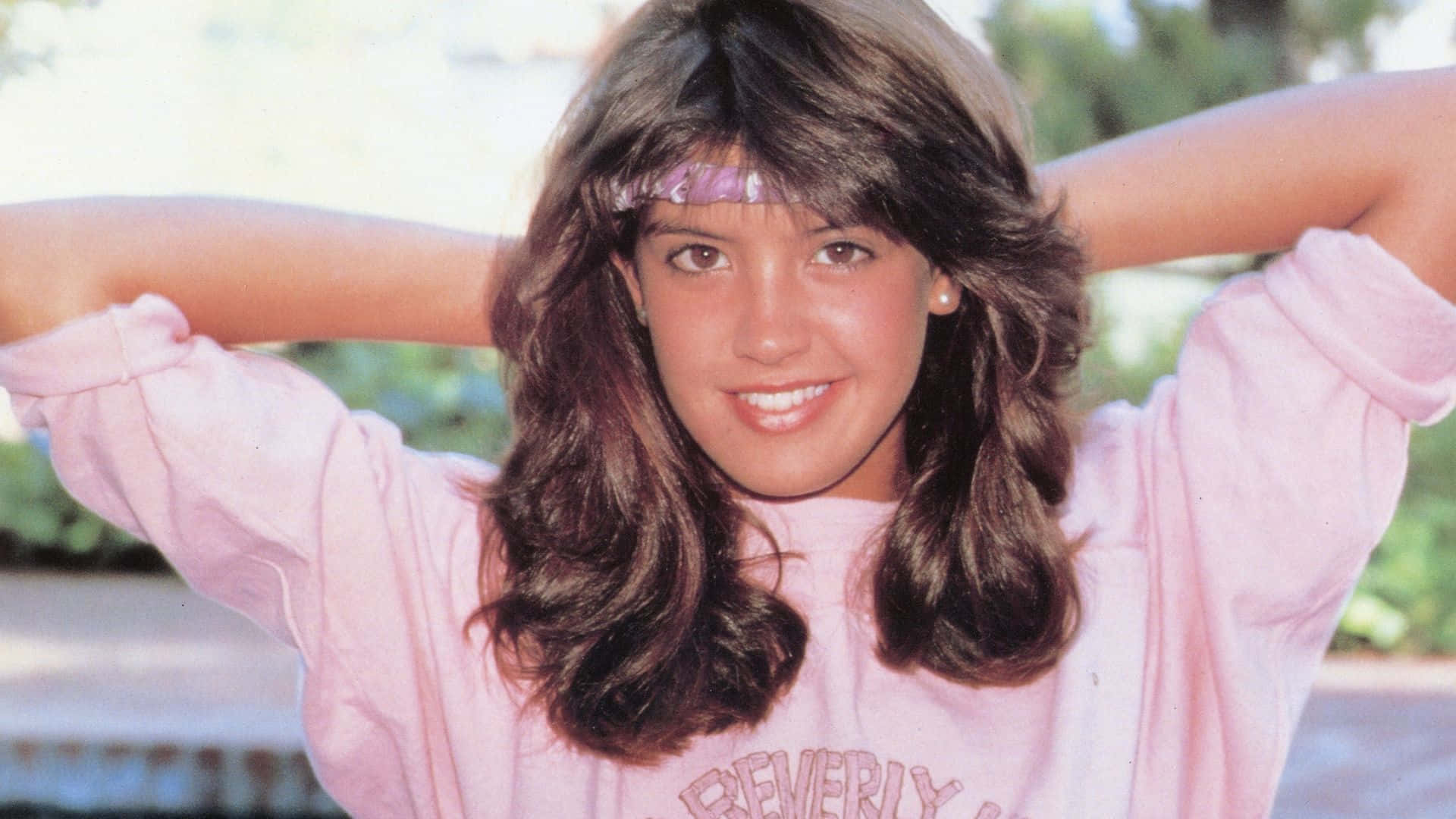 Phoebe Cates 桌布 壁紙 — 一位微笑的年輕女性，擁有豐盈的棕色頭髮和粉紅色頭帶，穿著粉紅色運動衫，舉起雙臂在戶外擺姿勢。