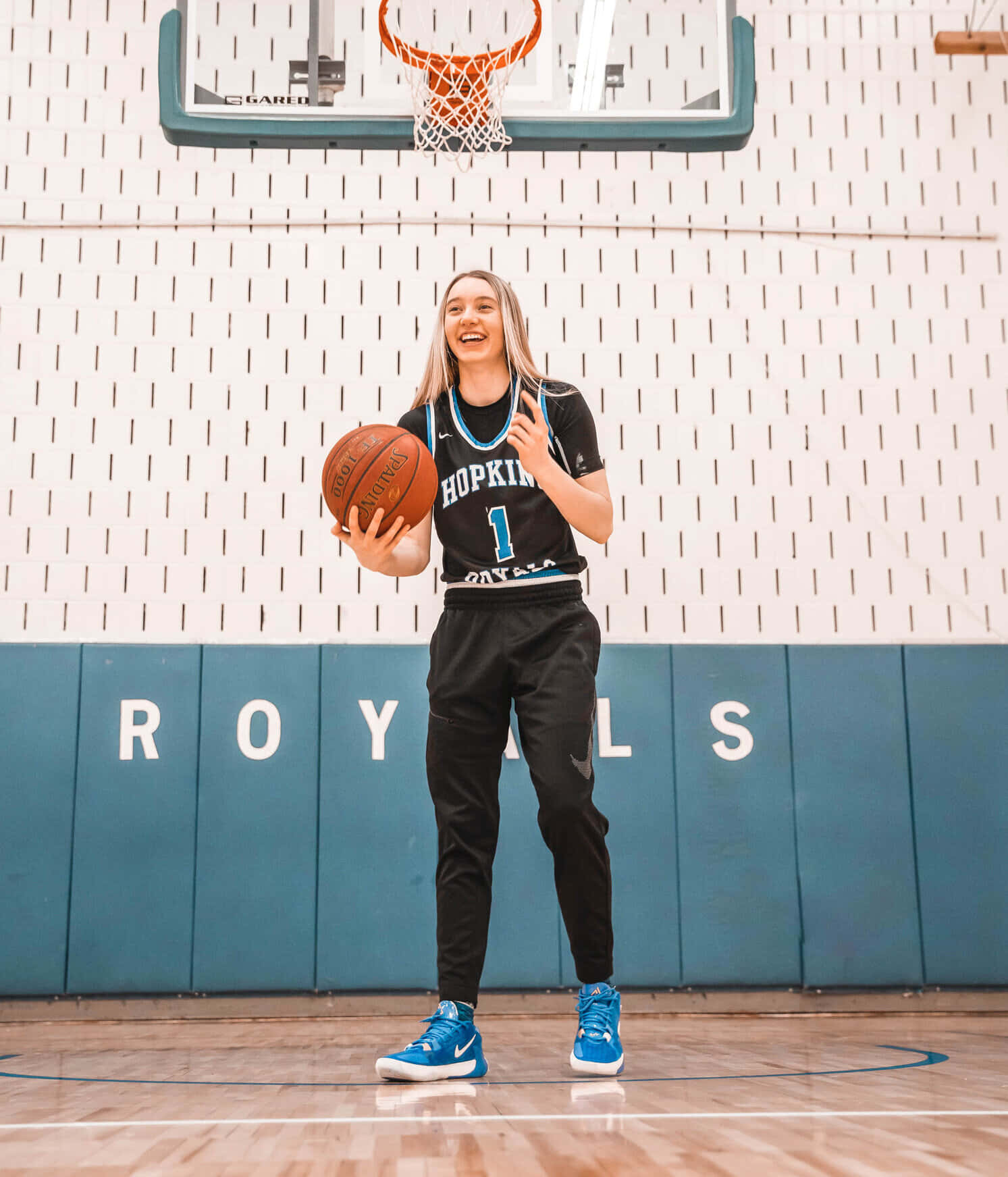  壁紙 — 一位微笑的女籃球員身穿黑色'Hopkins'球衣，在拋光木地板的球場上於籃框下持球。
