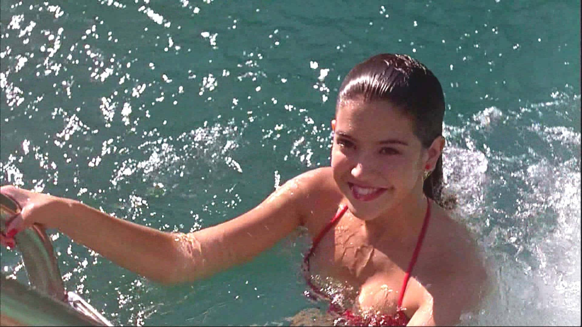 Phoebe Cates 桌布 壁紙 — 一位微笑的女士身穿紅色比基尼，從碧綠色的水中浮出，手握船欄杆。