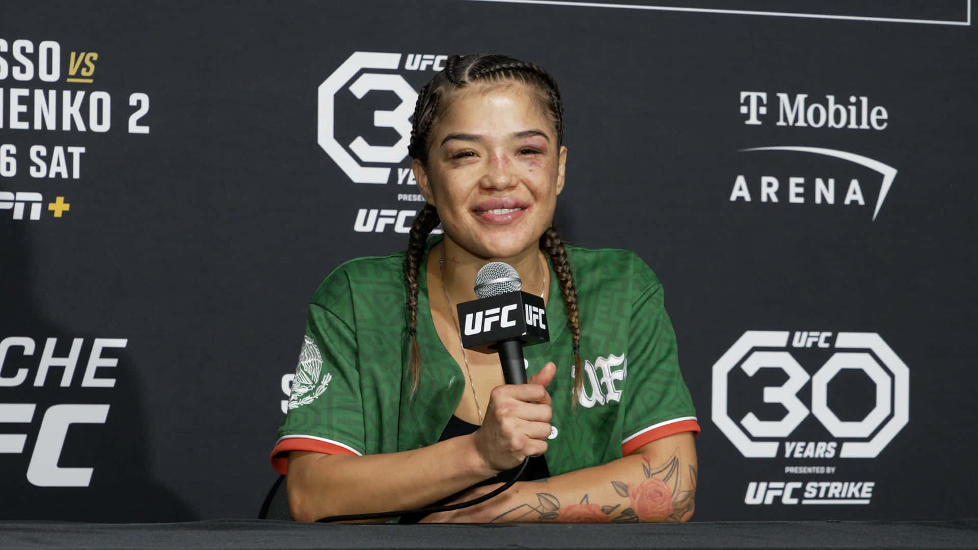  壁紙 — 一位微笑的女性UFC拳手身穿綠色球衣，在記者會上手持麥克風，背景有UFC 30標誌和T-Mobile Arena品牌標識。