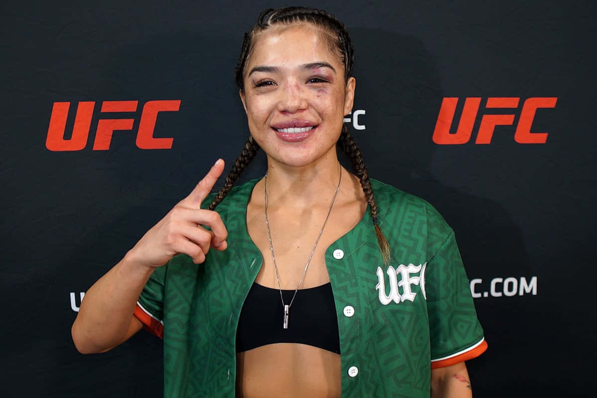  壁紙 — 一位微笑的UFC拳手，編織頭髮，帶有可見瘀傷，自信地指向鏡頭，背景為品牌背景。