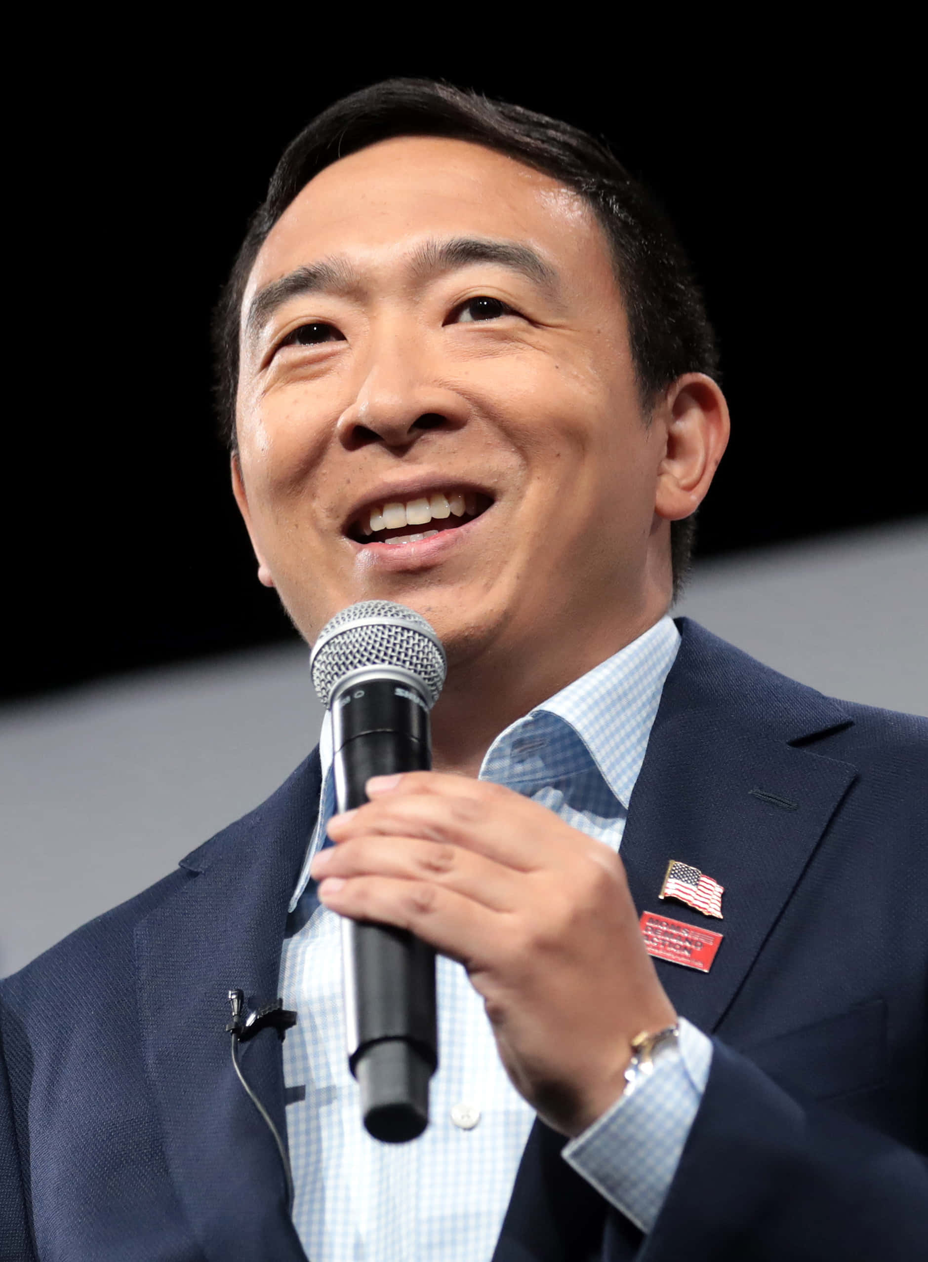 美國 桌布 壁紙 — 一位微笑的西裝男士在黑暗背景的活動中手持麥克風發言。