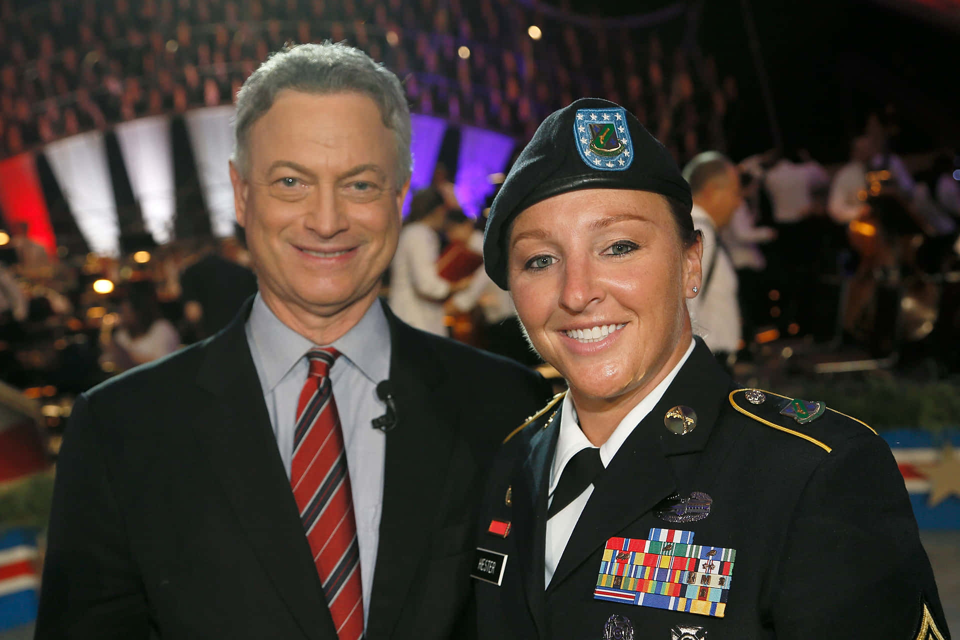  壁紙 — 一位微笑的西裝男子和一位穿著裝飾軍服的女子在正式活動中一起擺姿勢，背景是模糊的觀眾。