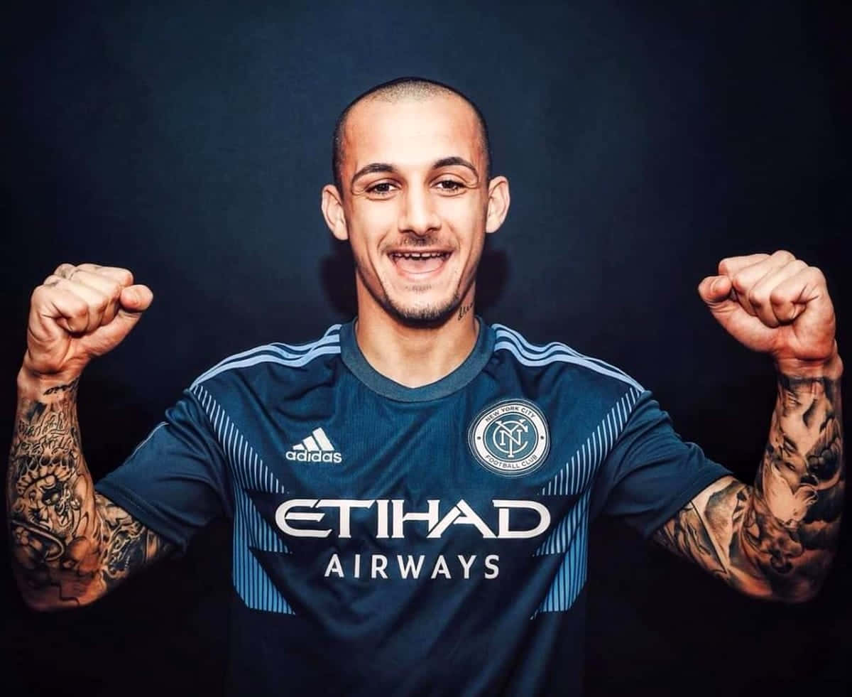  壁紙 — 一位微笑的足球運動員身穿深藍色NYCFC球衣，在黑色背景前握緊雙拳，露出手臂上複雜的紋身。