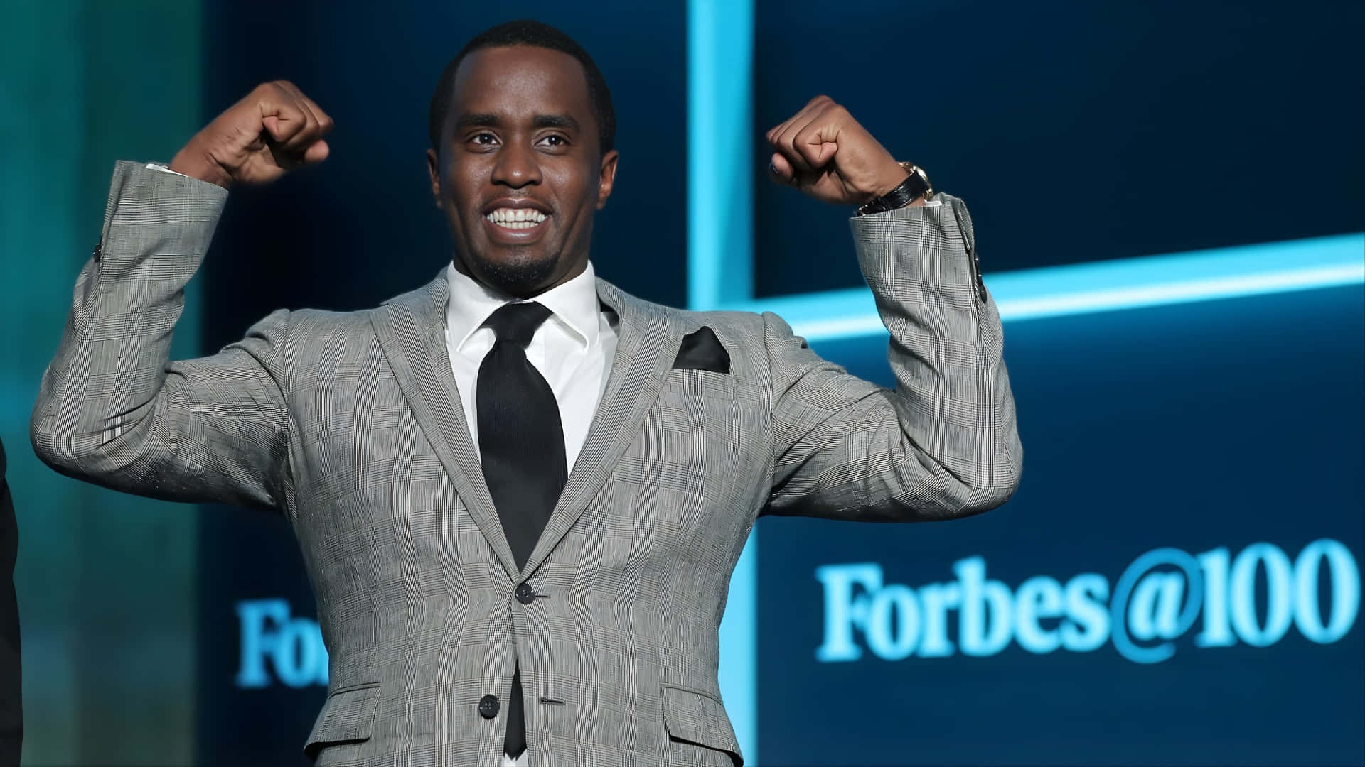  壁紙 — 一位微笑的男子身穿灰色西裝，在藍色 Forbes@100 背景的舞台上握緊雙拳。