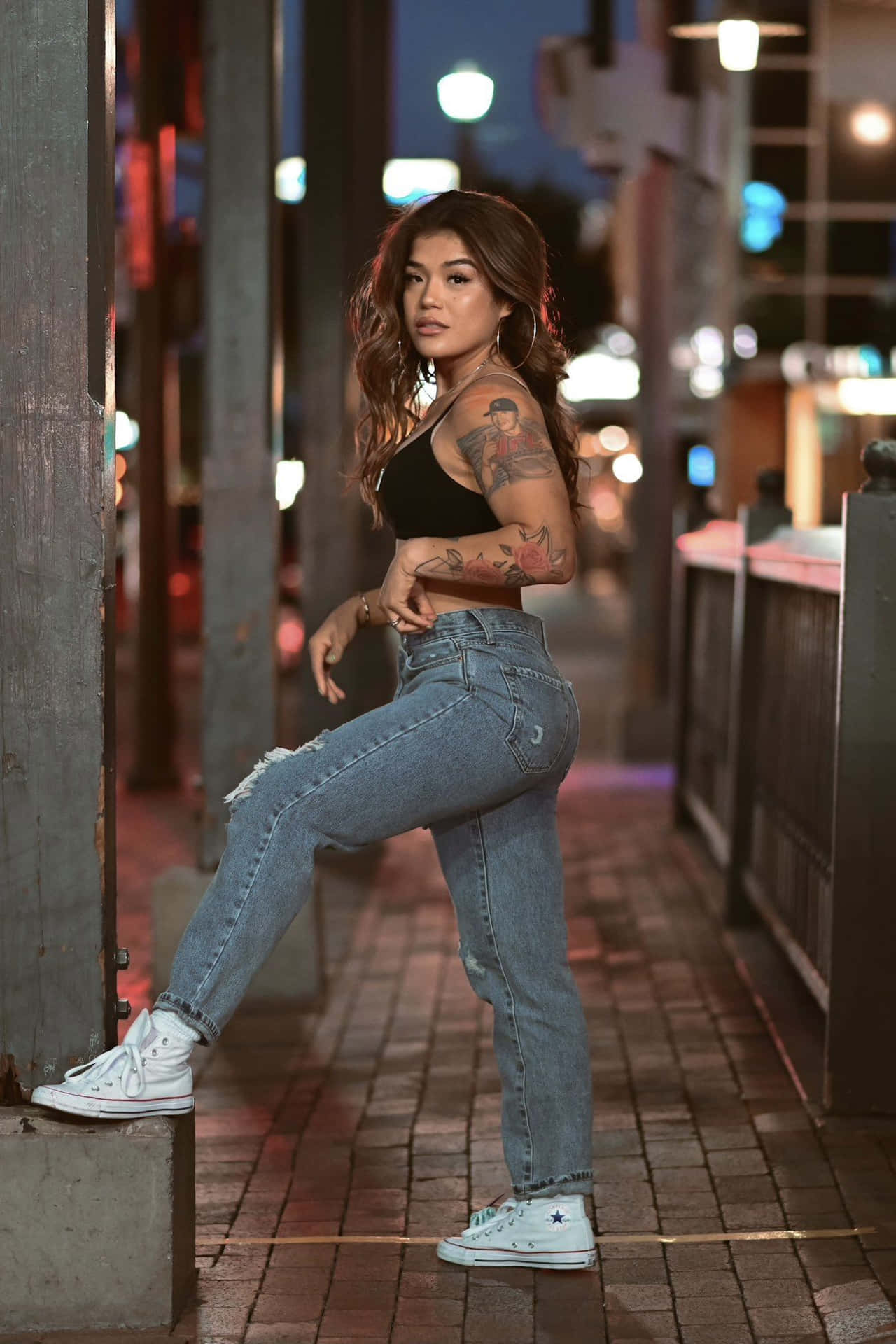  壁紙 — 一位紋身女子穿著破洞牛仔褲和白色運動鞋，在夜晚的城市人行道上自信地擺姿勢，身後是模糊的霓虹燈。