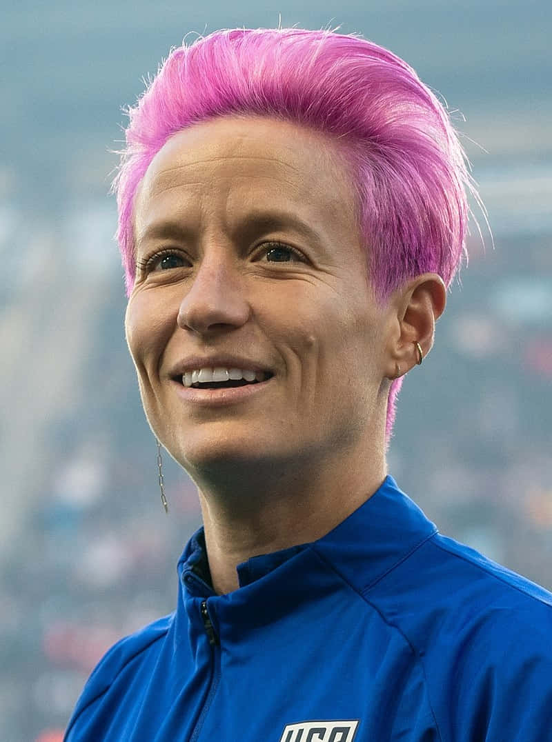  壁紙 — 一位笑容滿面的女士，留著充滿活力的粉紅色短髮，穿著藍色美國運動外套，背景是模糊的體育場人群。