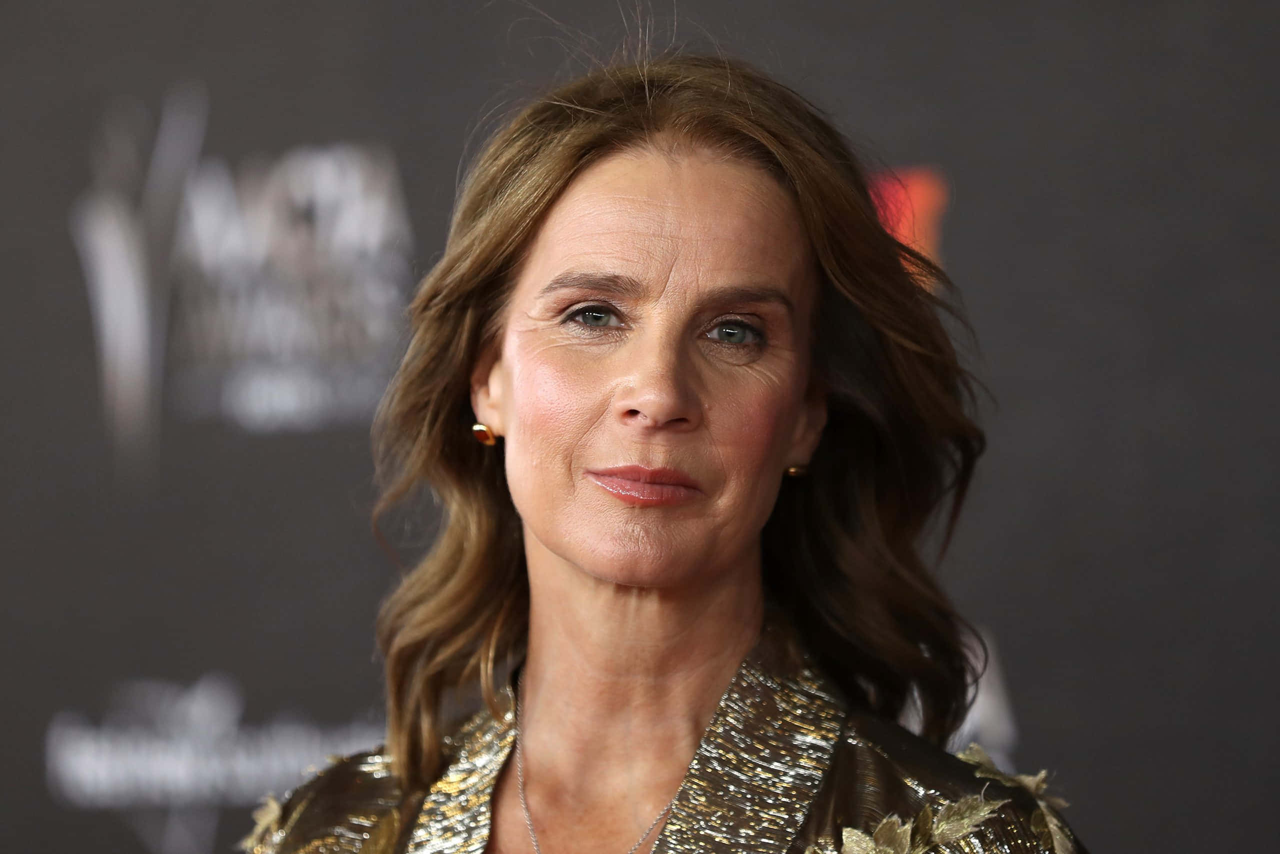 Rachel Griffiths 桌布 壁紙 — 一位擁有波浪棕色頭髮的女性在紅地毯活動中穿著閃閃發光的金色外套的 close-up 肖像。