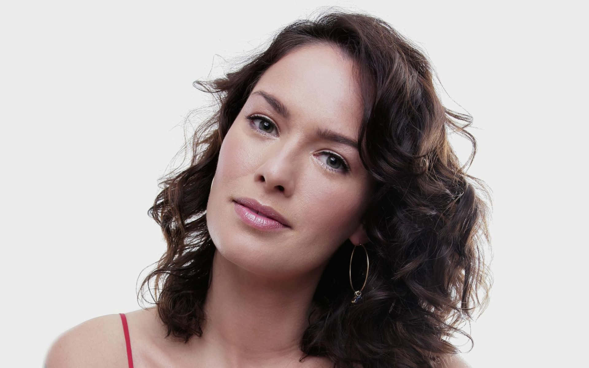 Lena Headey 桌布 壁紙 — 一位擁有波浪棕色頭髮和圓環耳環的女性，在純白背景前溫柔地凝視。