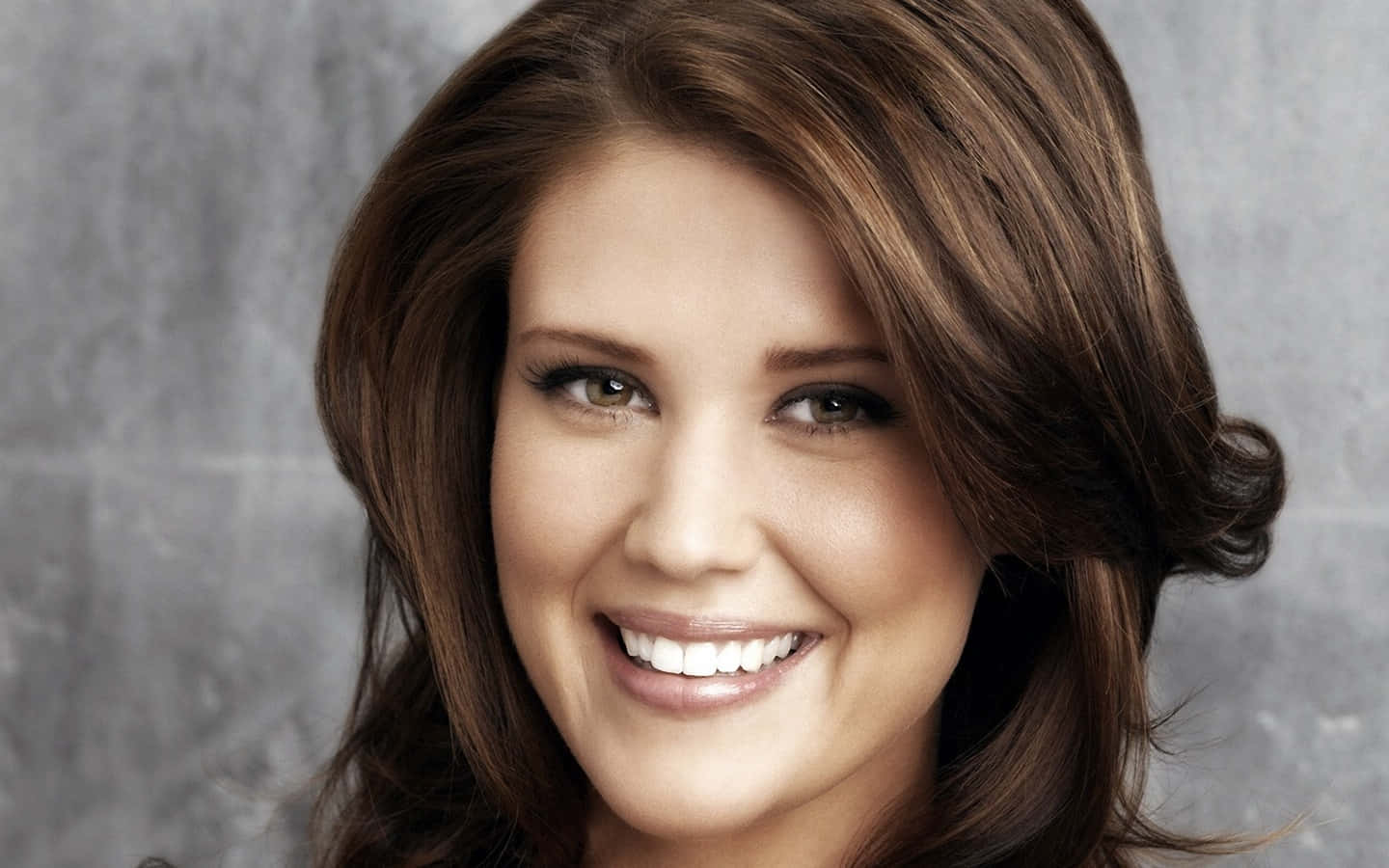 Sarah Lancaster 桌布 壁紙 — 一位擁有棕色頭髮的燦爛女性，在帶有紋理的灰色背景前溫暖地微笑。