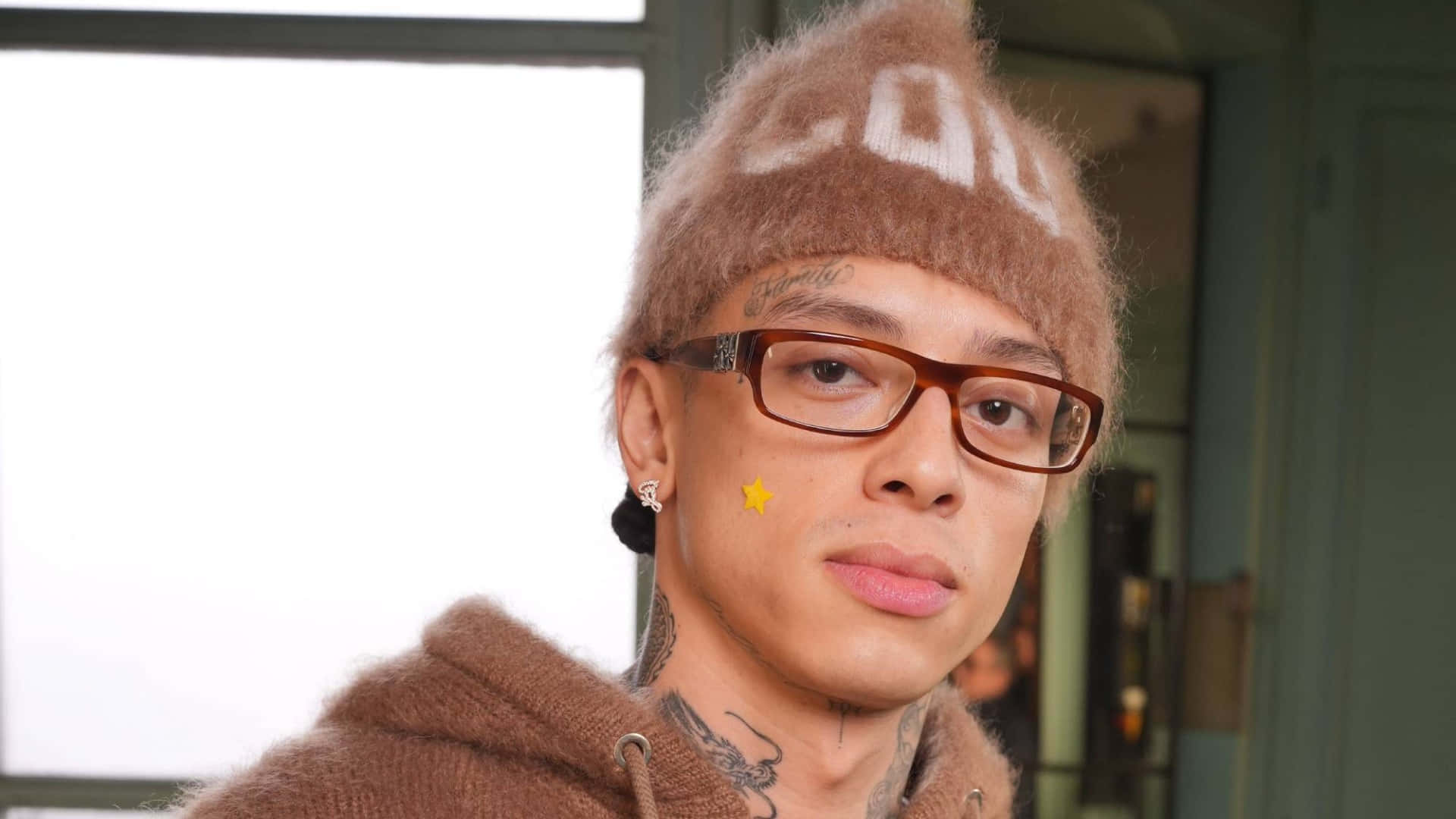  壁紙 — 一位有紋身的年輕男子戴著毛絨棕色冷帽和眼鏡，直視鏡頭。