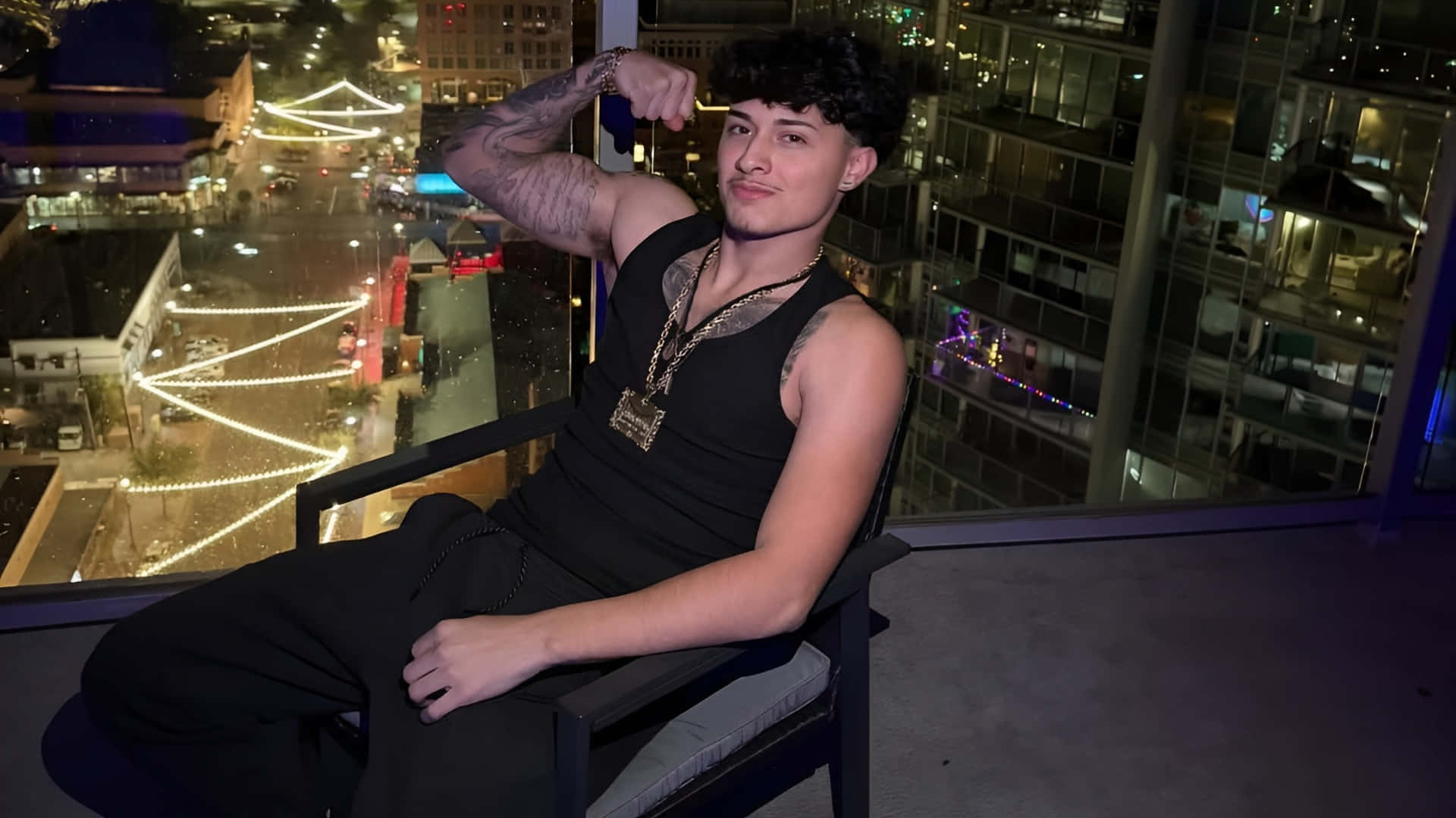  壁紙 — 一位有紋身的年輕男子坐在高層大廈陽台的椅子上，對著夜晚充滿活力的城市景觀彎曲二頭肌。