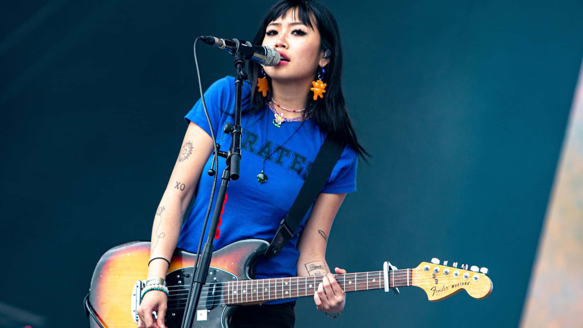  壁紙 — 一位有紋身的女音樂家在藍色燈光下在舞台上彈奏旭日色電結他並對著咪高峰唱歌。