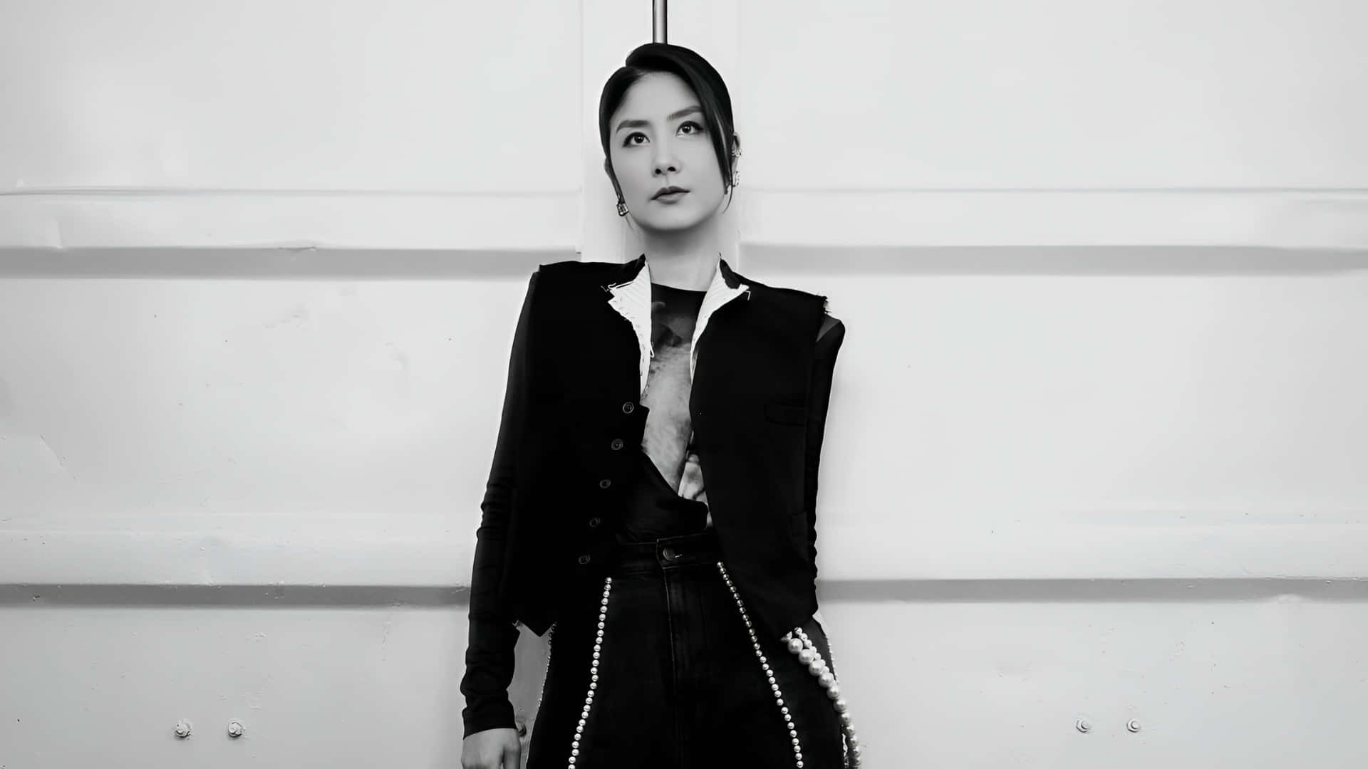  壁紙 — 一位優雅女士身穿黑色西裝外套和珍珠裝飾褲子，站在單色極簡白色牆壁前。