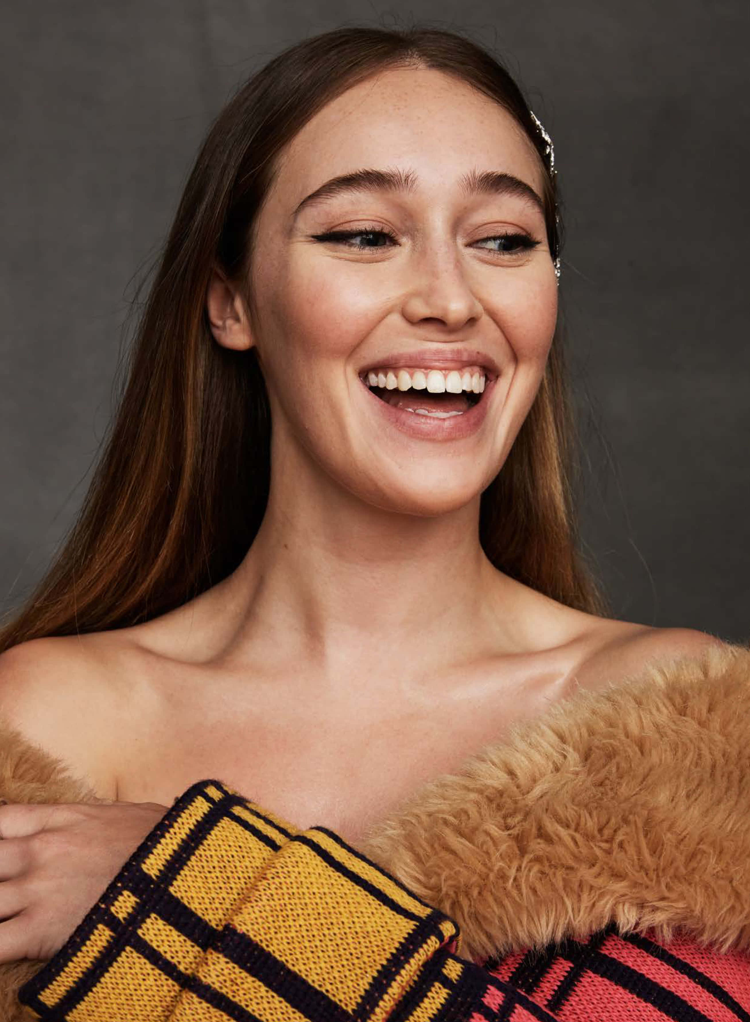 alycia debnam carey 桌布 壁紙 — 一位長著棕色長髮的快樂女性，在灰色背景前，抱著一條帶毛邊的彩色格仔毯，笑得非常燦爛。