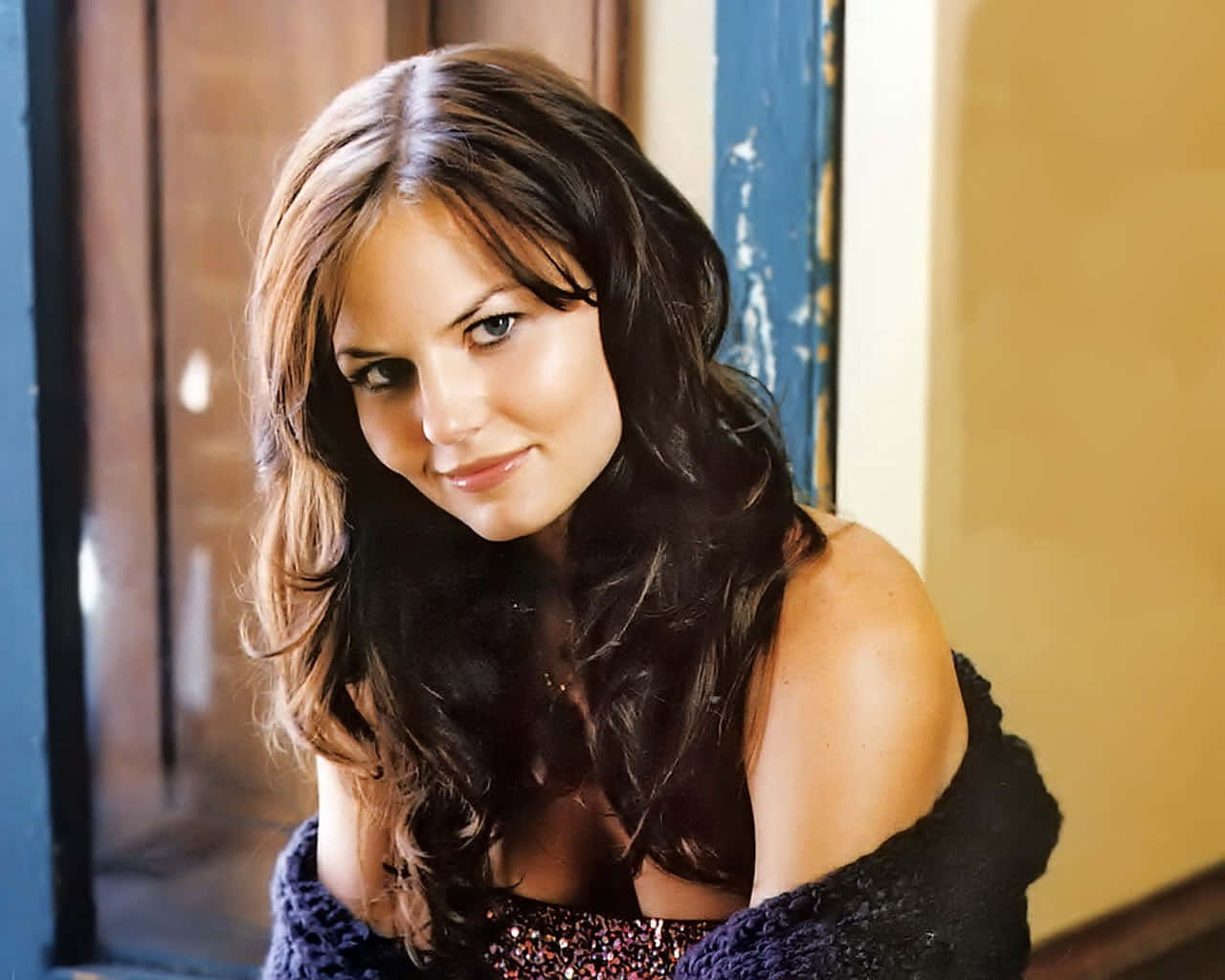 jennifer morrison 桌布 壁紙 — 一位長著深色波浪長髮的女性，輕輕微笑著靠在一個風化的藍色木門框上。