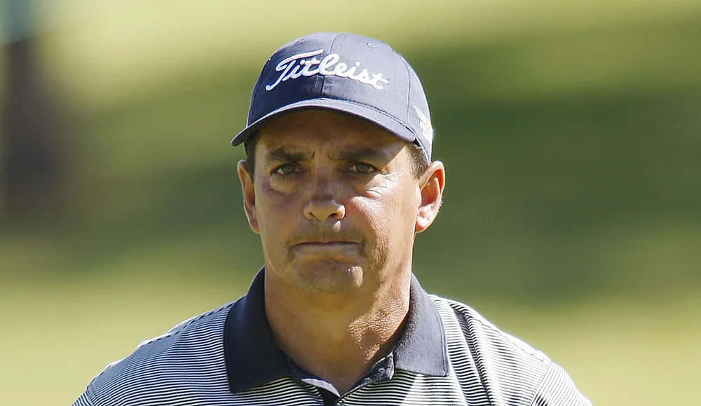  壁紙 — 一位專注的高爾夫球手戴著 Titleist 帽子和條紋 Polo 衫，站在陽光明媚的高爾夫球場上，身後是模糊的綠化。