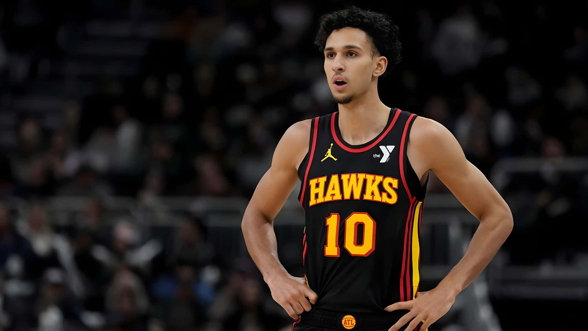  壁紙 — 一位專注的籃球運動員身穿黑色Hawks球衣，號碼10，雙手叉腰站在球場上。