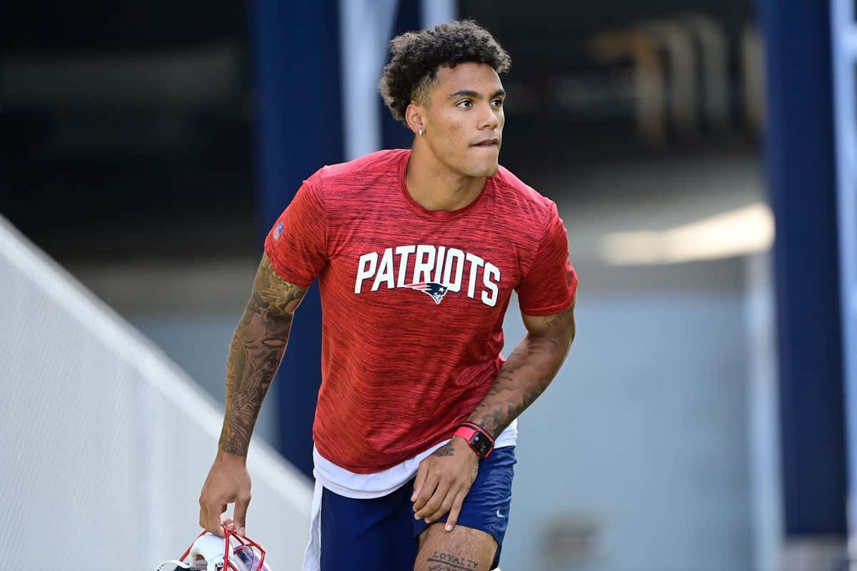 Christian Gonzalez 桌布 壁紙 — 一位專注的美式足球員穿著紅色Patriots球衣，在訓練期間拿著頭盔行走。