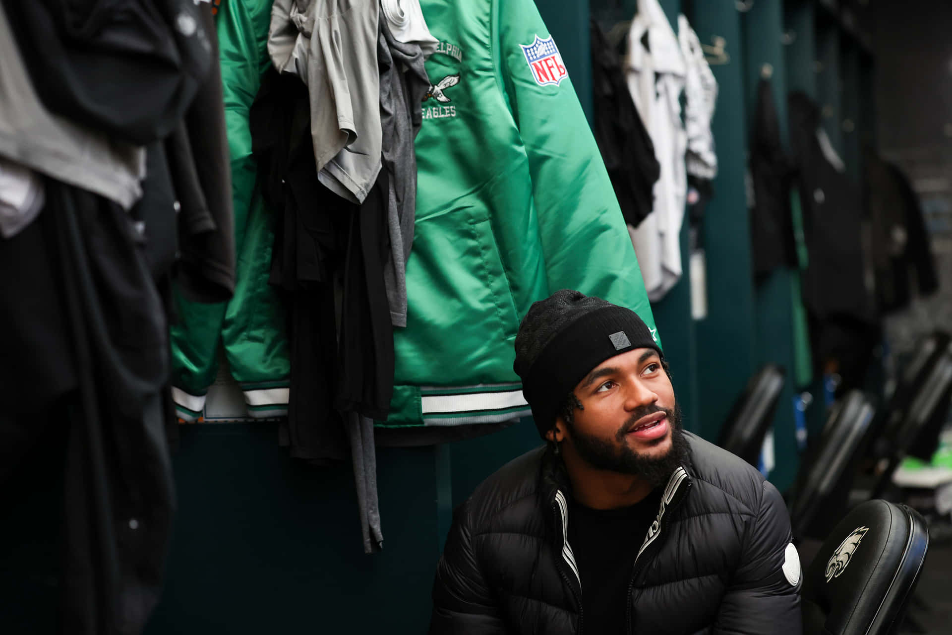  壁紙 — 一位專注的NFL球員戴著黑色冷帽和羽絨外套，坐在被掛著的Eagles裝備包圍的更衣室裡。