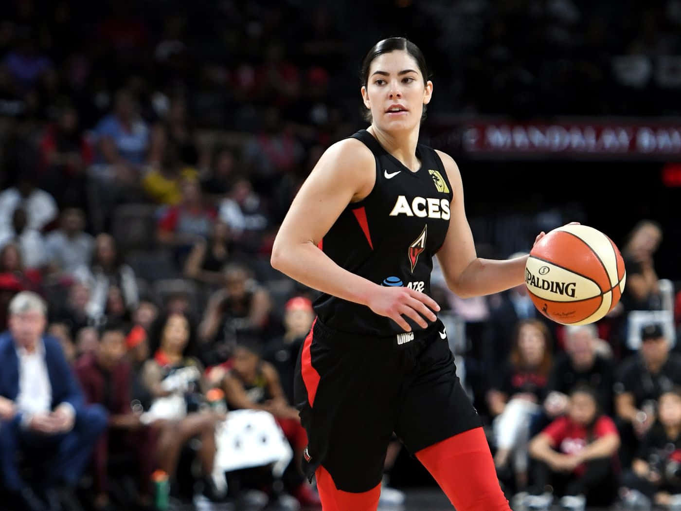  壁紙 — 一位專注的女籃球員身穿黑色和紅色的「ACES」球衣，在場館燈光下進行激烈比賽時運著Spalding籃球。