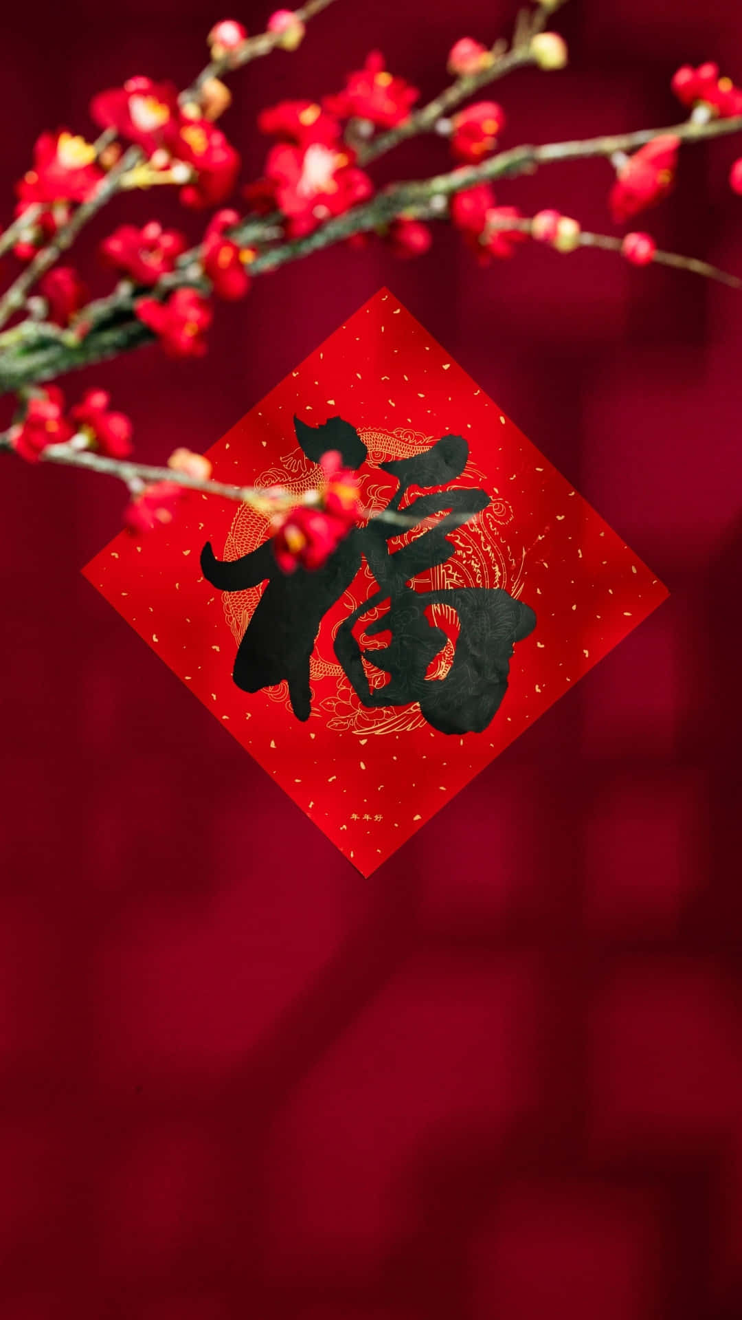  壁紙 — 一張紅色菱形紙，上面有黑色漢字福，掛在深紅色背景上，背景點綴著盛開的紅色梅花。