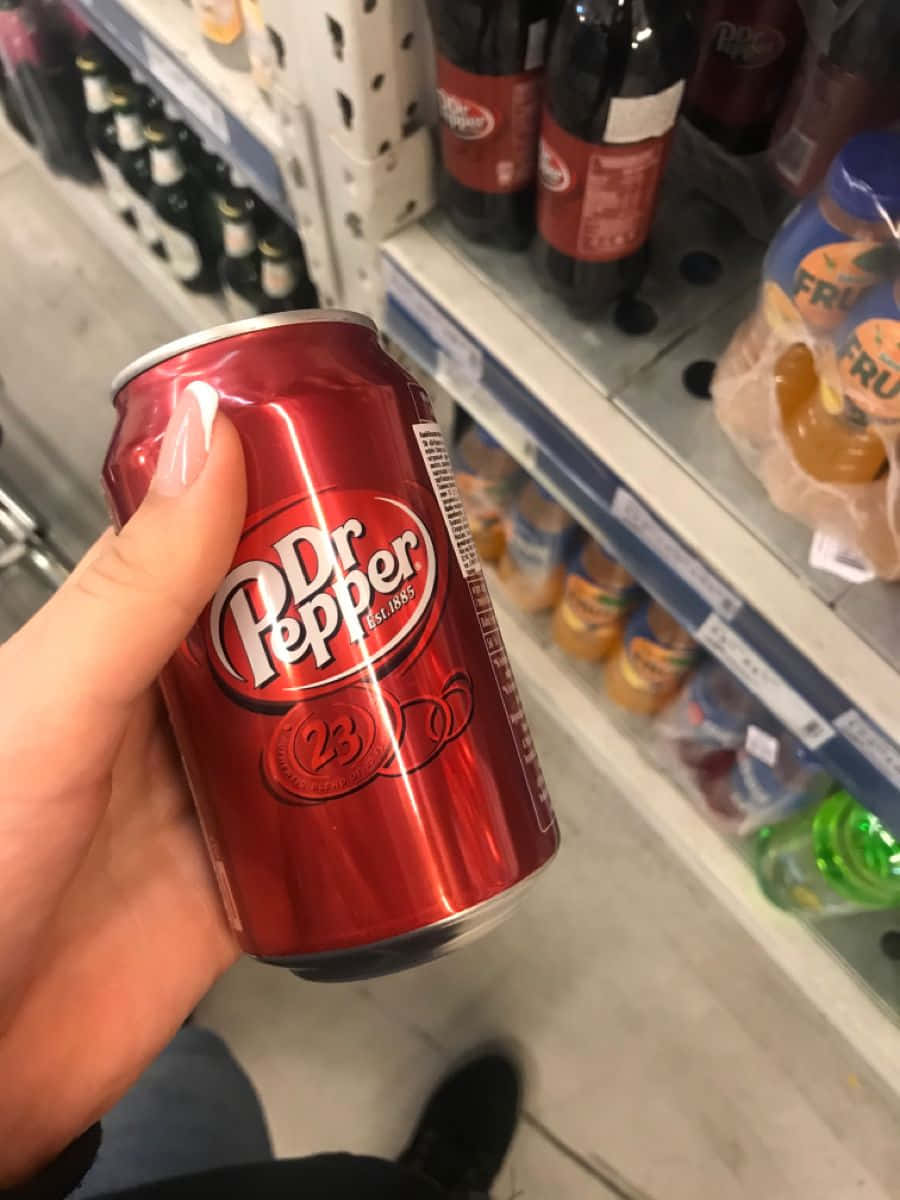  壁紙 — 一隻手在光線充足的雜貨店通道中拿著一個閃亮的紅色 Dr Pepper 23 罐頭，後面是擺滿飲料的貨架。