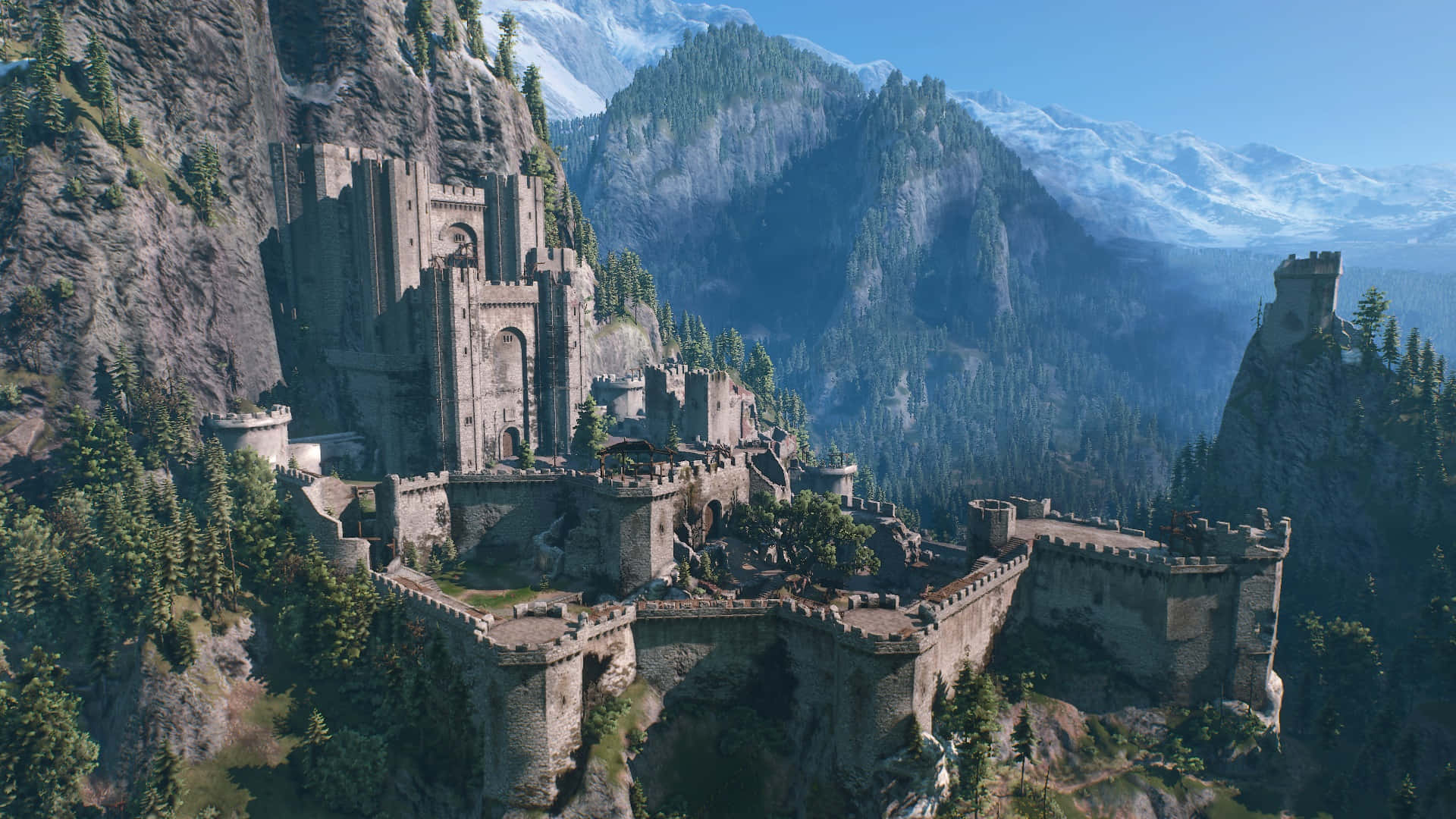 Kaer Morhen 桌布 壁紙 — 一座宏偉的中世紀城堡坐落在懸崖上，俯瞰著山谷，山谷中有積雪覆蓋的山脈和茂密的森林，天空湛藍清澈。