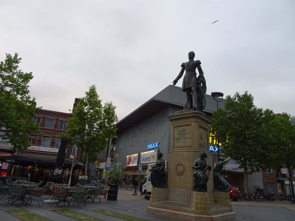 Tilburg 桌布 壁紙 — 一座歷史人物的銅像高高矗立在城市廣場的石基座上，周圍環繞著樹木和現代建築，天空多雲。
