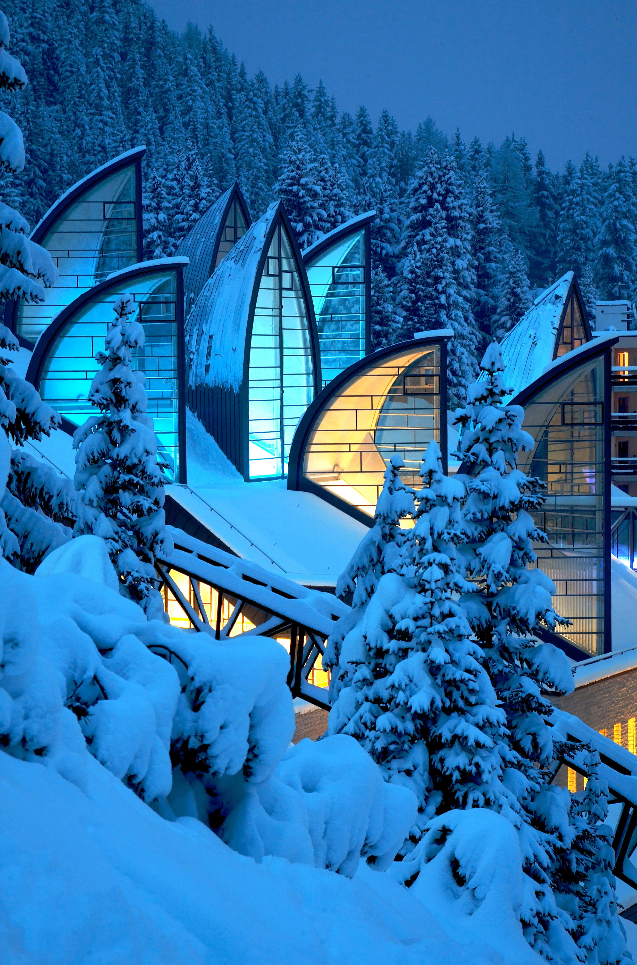 Switzerland 桌布 壁紙 — 一座現代玻璃建築，擁有帆狀屋頂，在暮色中對著雪林溫暖地發光。