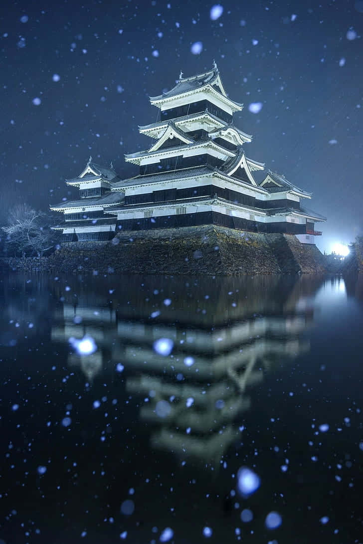  壁紙 — 一座雄偉的日本城堡在夜晚被照亮，其倒影在平靜的水面上閃爍，雪花在黑暗的天空中飄落。