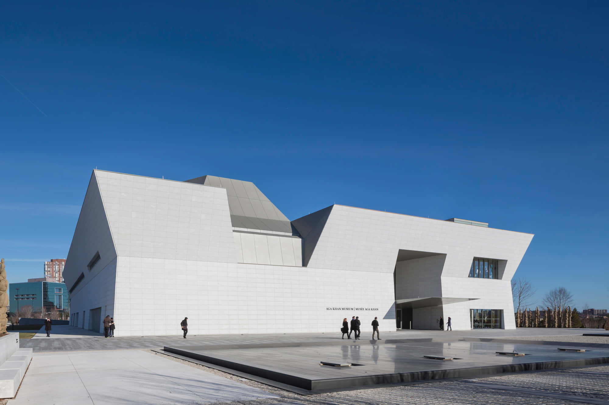 Aga Khan Museum 桌布 壁紙 — 一座引人注目的現代白色建築結構，帶有銳利的幾何角度，矗立在晴朗的藍天下，廣場上有人們在散步。