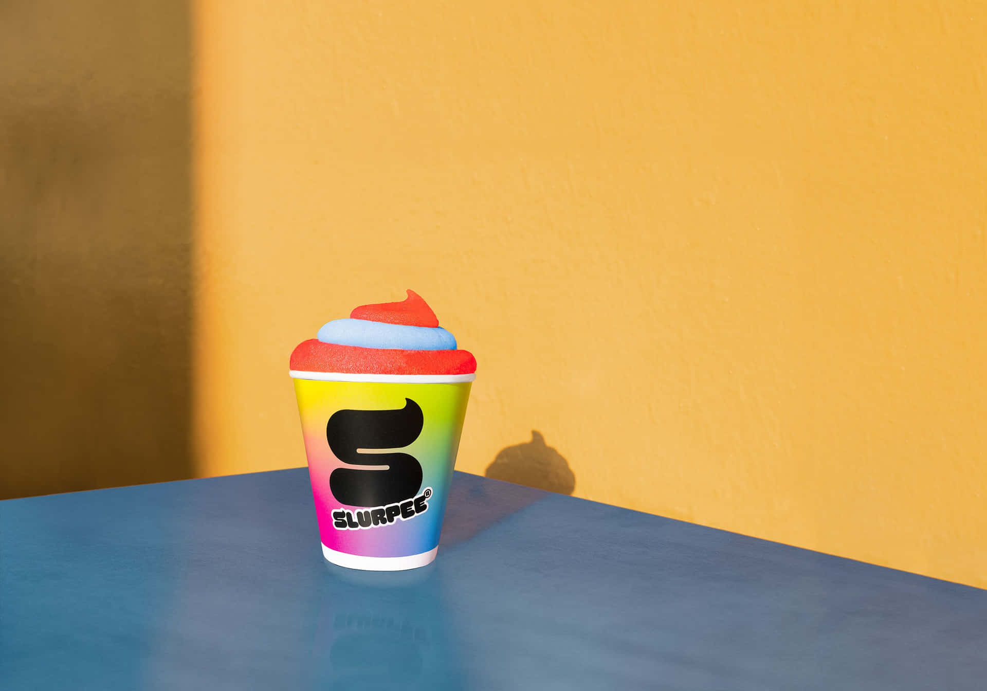  壁紙 — 一個充滿活力的彩虹 Slurpee 杯，帶有紅色和藍色漩渦，放在藍色桌子上，背景是溫暖的黃色牆壁。
