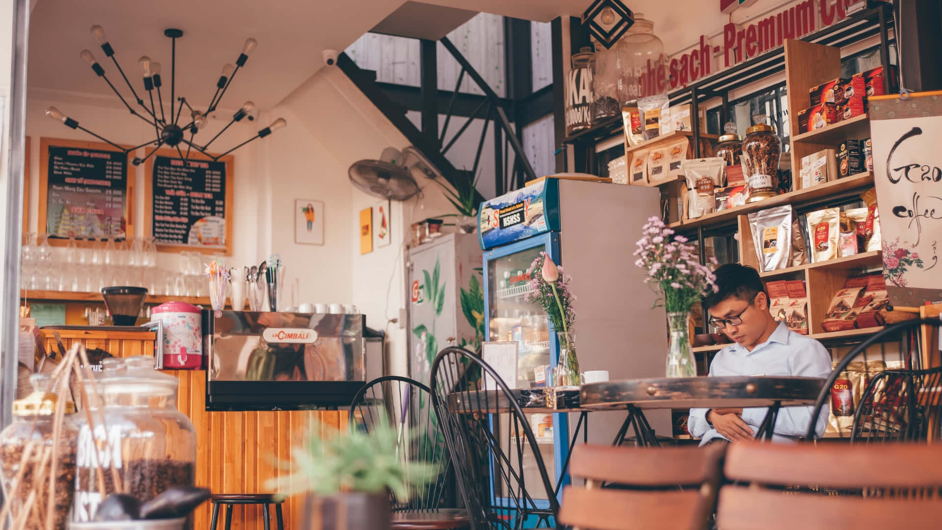 Coffee Shop 桌布 壁紙 — 一個戴眼鏡的年輕男子坐在舒適咖啡館的木桌旁，周圍是咖啡豆架和復古裝飾。