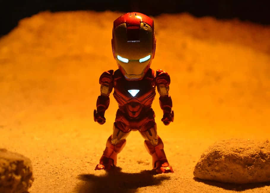 Iron Man Bobbleheads 桌布 壁紙 — 一個發光的紅色和金色鐵甲奇俠人偶在溫暖的橙色燈光下英勇地站在沙地上。