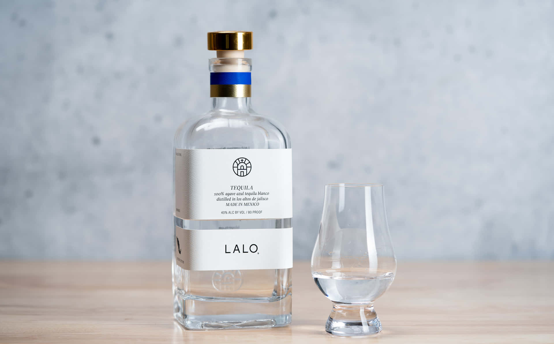  壁紙 — 一個透明嘅Lalo龍舌蘭酒樽，金色蓋同藍色帶，放喺木質表面上，旁邊有個空嘅品酒杯，背景係有紋理嘅灰色牆。