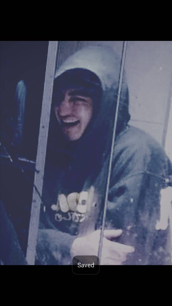 Joji 閃耀笑容海報 壁紙 — 一個穿著連帽外套的人喺一塊有明顯污漬同反射嘅玻璃後面開心咁笑。