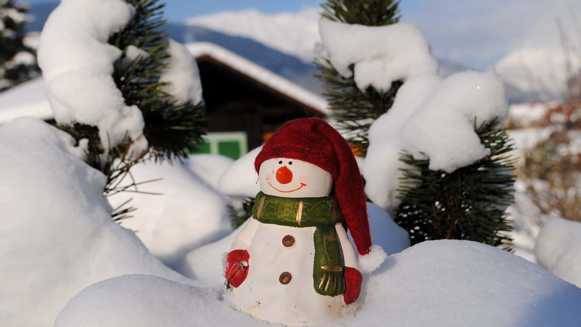  壁紙 — 一個戴紅帽、圍綠圍巾的快樂雪人站在厚雪中，旁邊是積雪的松樹，背景有一座木屋。