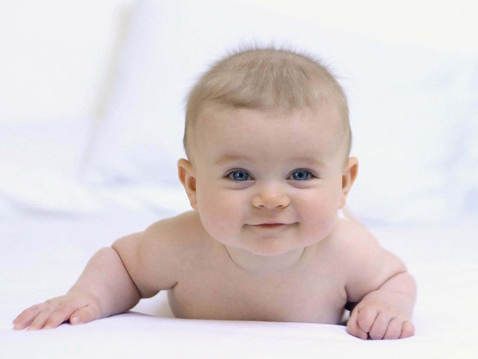  壁紙 — 一個開心的嬰兒，有著明亮的藍眼睛，躺在柔軟的白色表面上微笑。