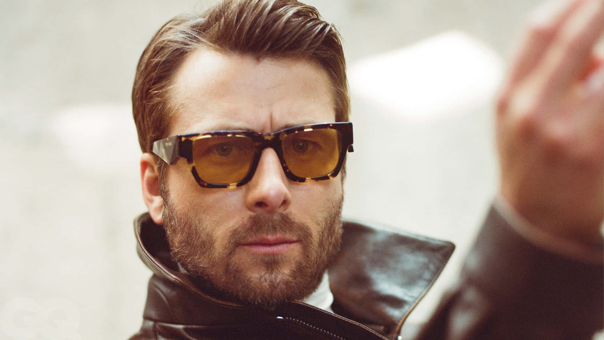  壁紙 — 一個留鬍鬚嘅男人戴住玳瑁色太陽眼鏡，著住皮褸，背景係模糊嘅城市。