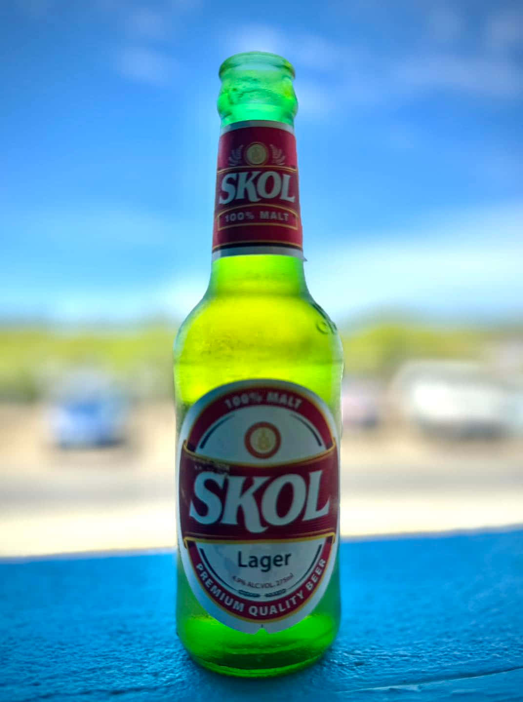  壁紙 — 一個鮮綠色嘅Skol啤酒樽放在藍色表面上，背景係明亮藍天同模糊嘅汽車。