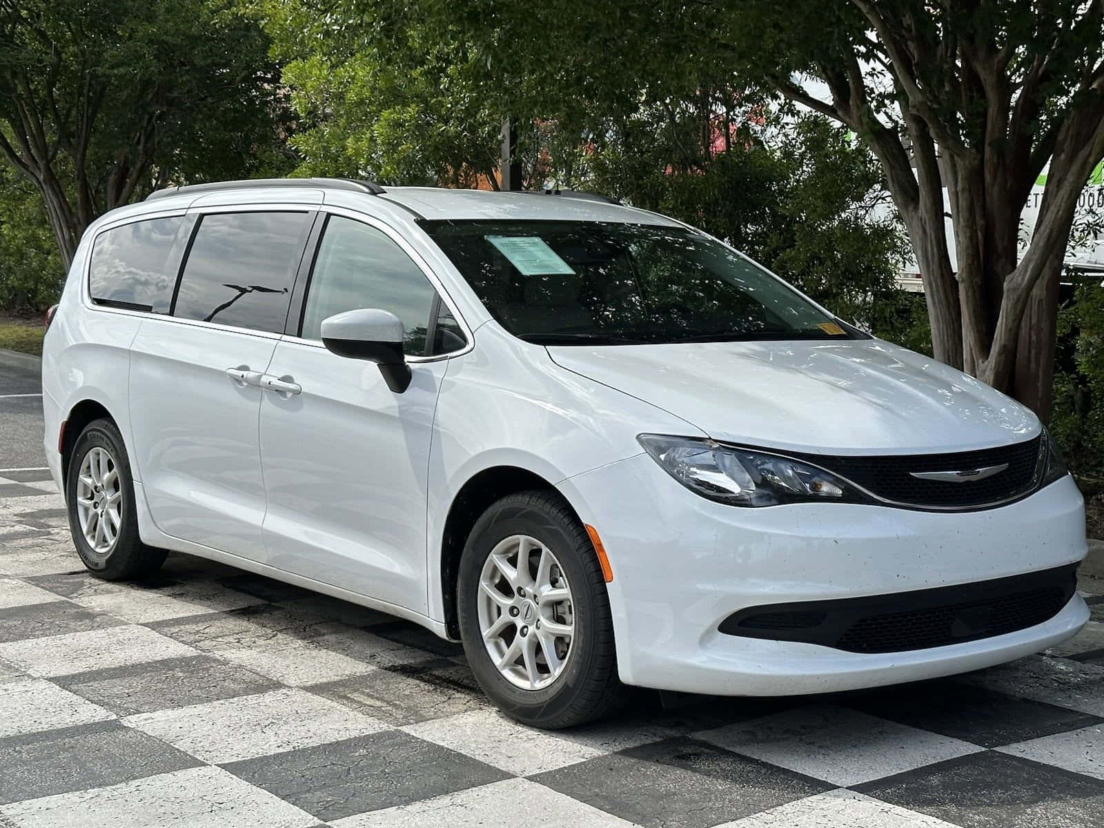 Chrysler Voyager 桌布 壁紙 — 一架純白色的 Chrysler Pacifica 小型貨車停泊在黑白格仔路面上，上方有綠葉成蔭的樹木。