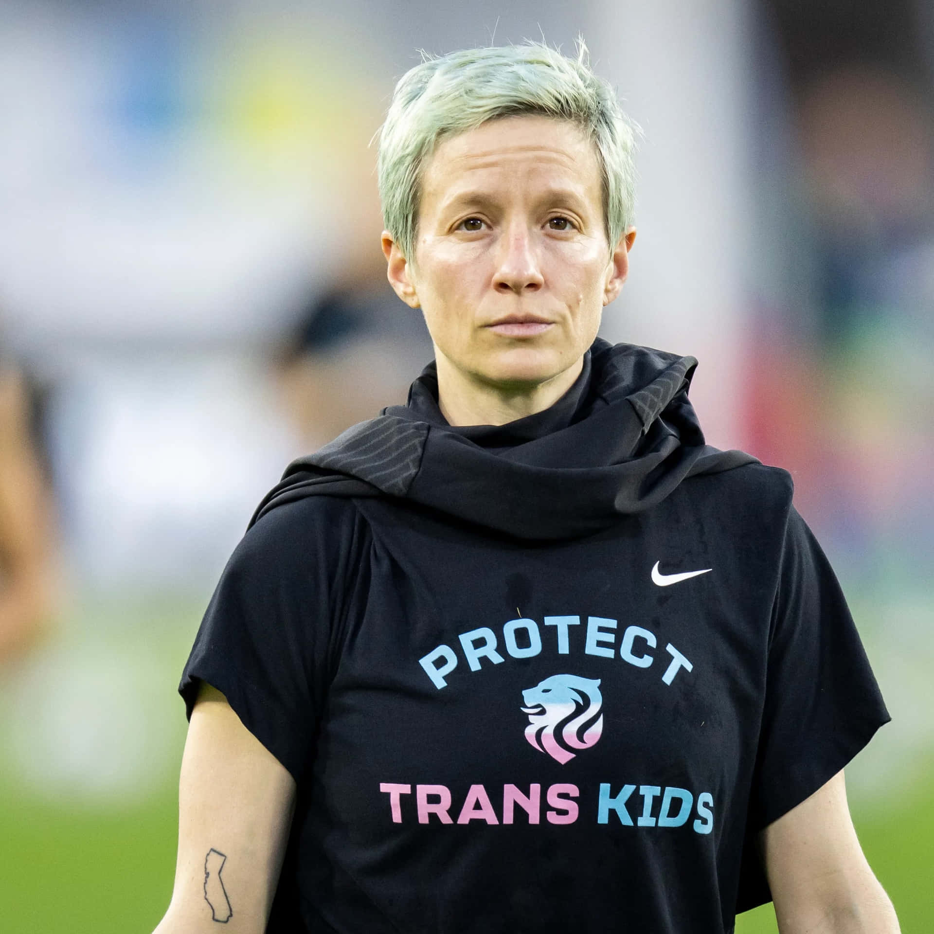  壁紙 — 一個短銀髮嘅人喺運動場着住一件黑色T恤，上面有'PROTECT TRANS KIDS'同獅子標誌。