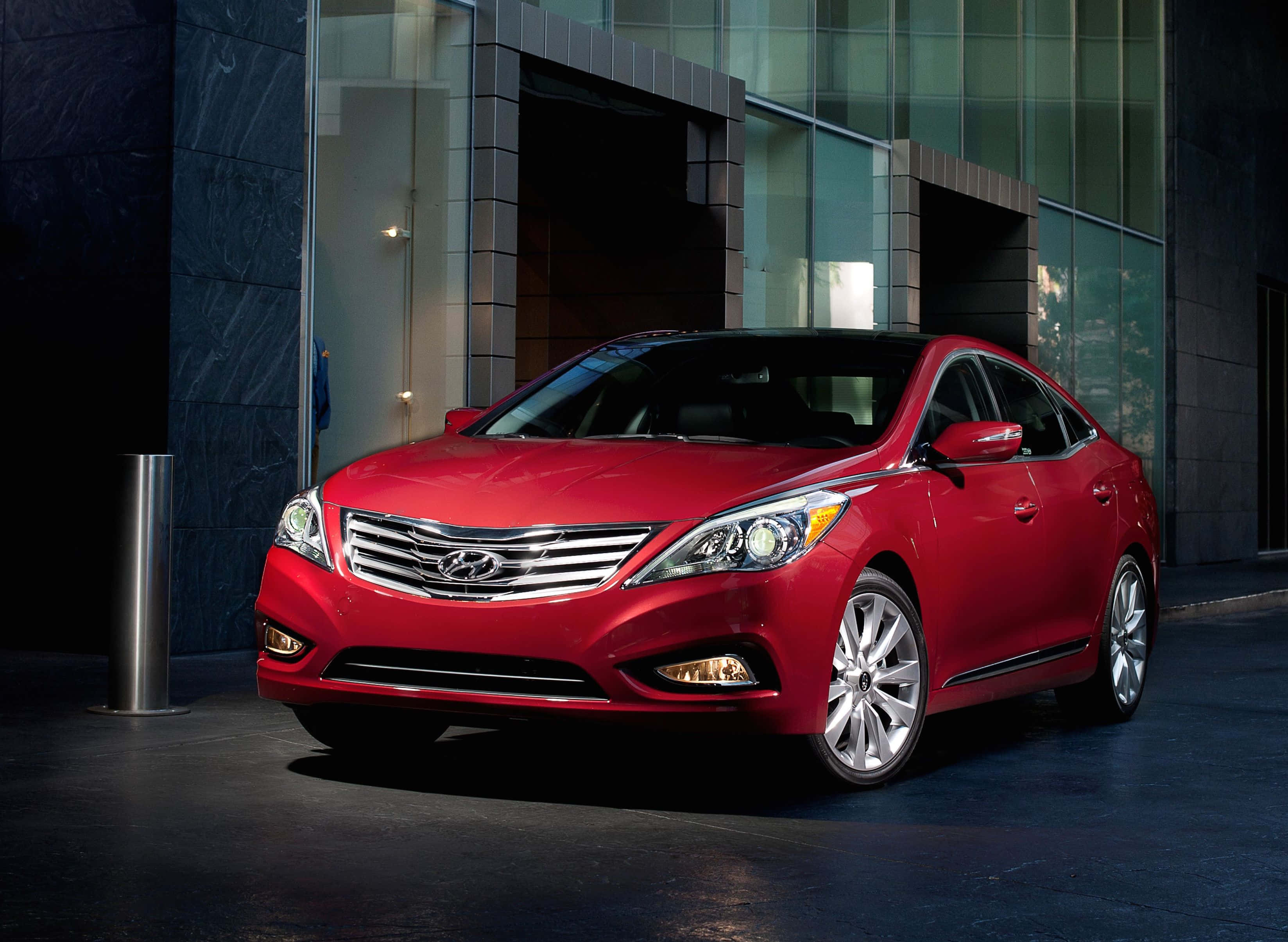 Hyundai Azera 桌布 壁紙 — 一輛光澤的紅色現代轎車在攝影棚燈光下閃耀，停在一棟現代玻璃和石材建築前。