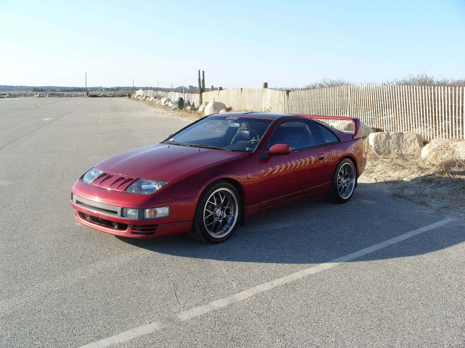 Nissan 300zx 桌布 壁紙 — 一架亮紅色日產300ZX跑車停泊在柏油路上，旁邊有木柵欄，天空湛藍清澈。
