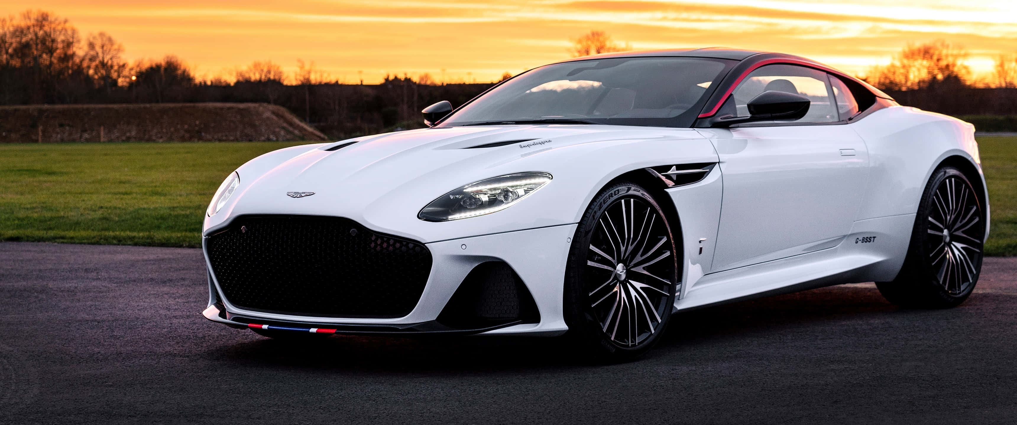 Aston Martin Dbs Superleggera 桌布 壁紙 — 一架流線型白色Aston Martin DBS Superleggera在日落時分停在瀝青路上，背後是金色天空和綠色田野。