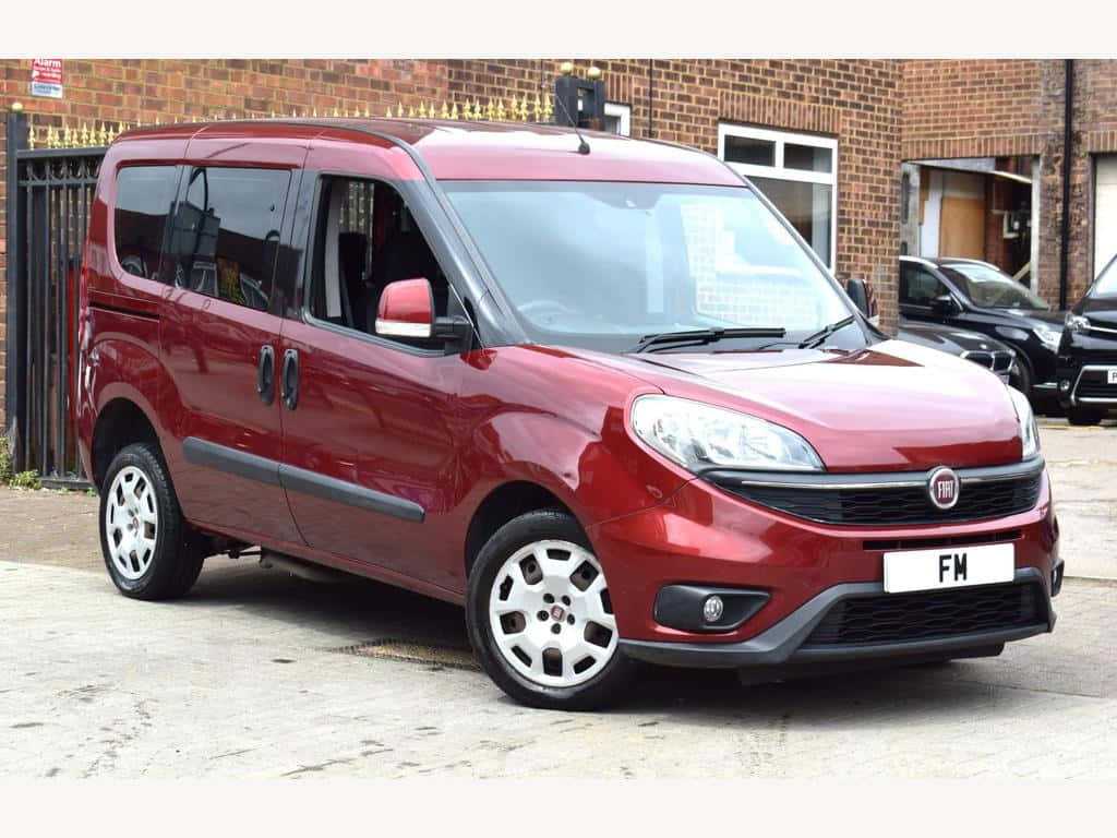  壁紙 — 一輛亮紅色的Fiat Fiorino客貨車停泊在磚砌建築物前的鋪砌場地上，建築物設有黑色欄杆。