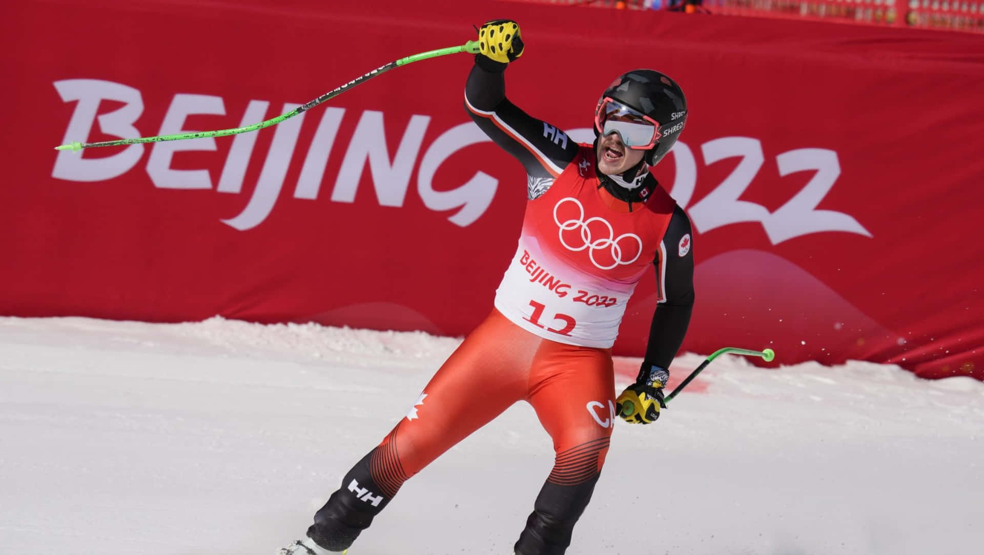  壁紙 — 一名身穿紅黑裝備的滑雪運動員在雪坡上慶祝勝利，身後有北京2022的橫幅。